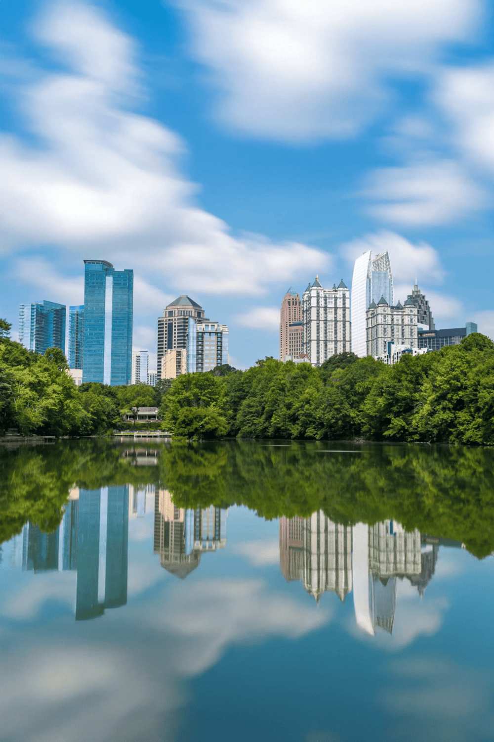 City skyline with modern buildings reflected in a calm lake, vibrant green trees, and blue sky with clouds.