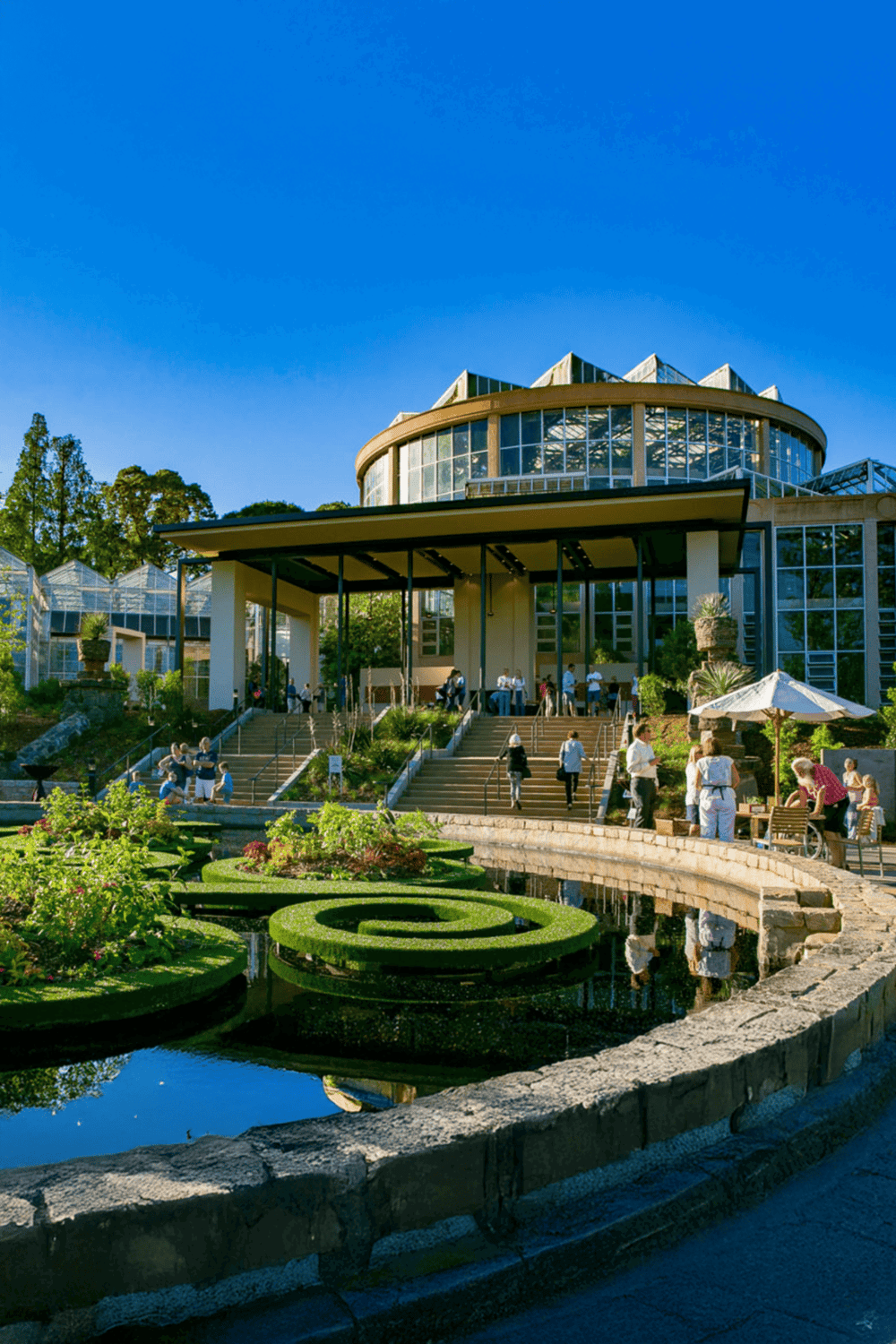 Modern botanical garden conservatory with glass architecture, lush greenery, and visitors enjoying outdoor spaces.