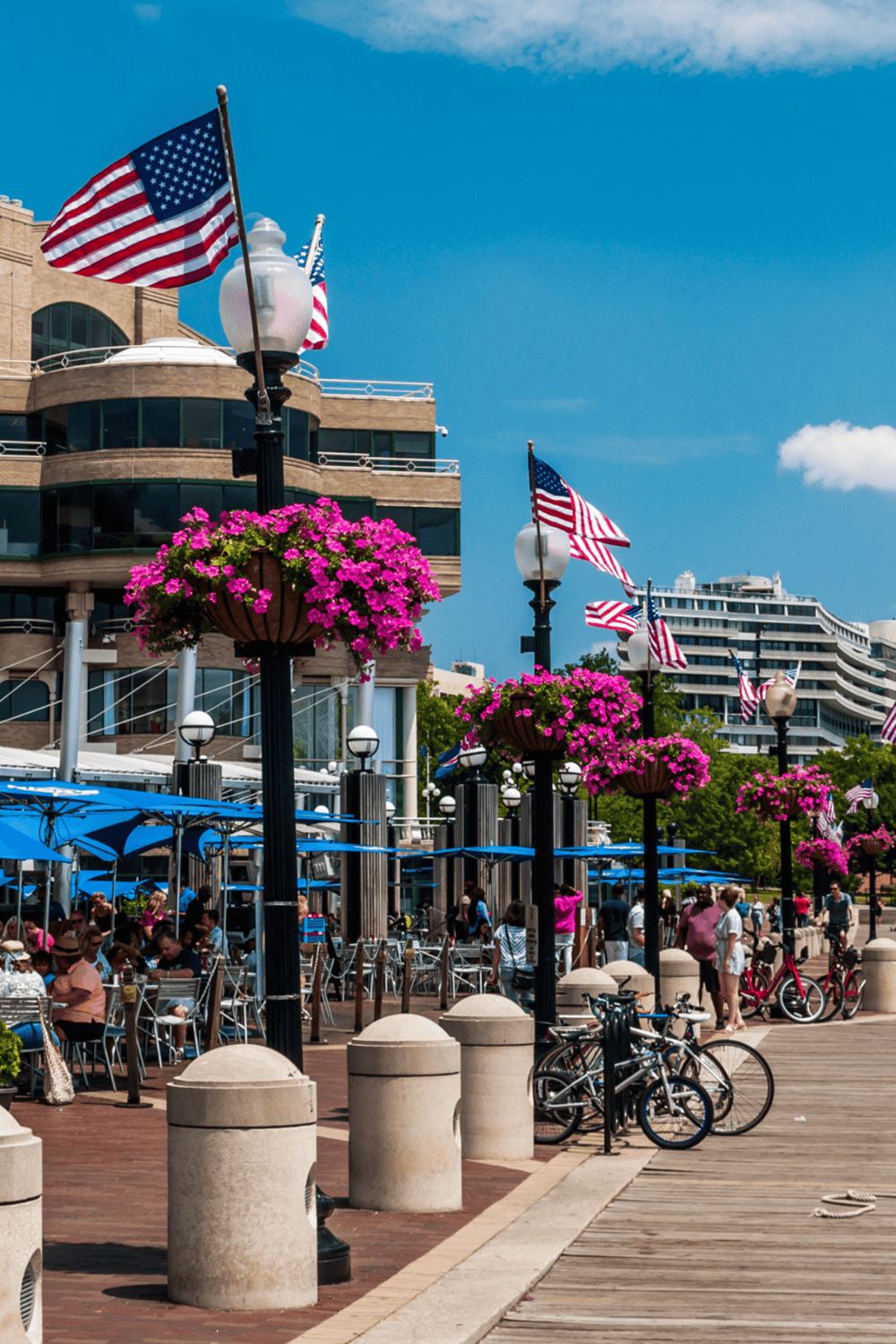 Flags on street lamps with pink flower baskets and outdoor café on a bustling waterfront promenade.