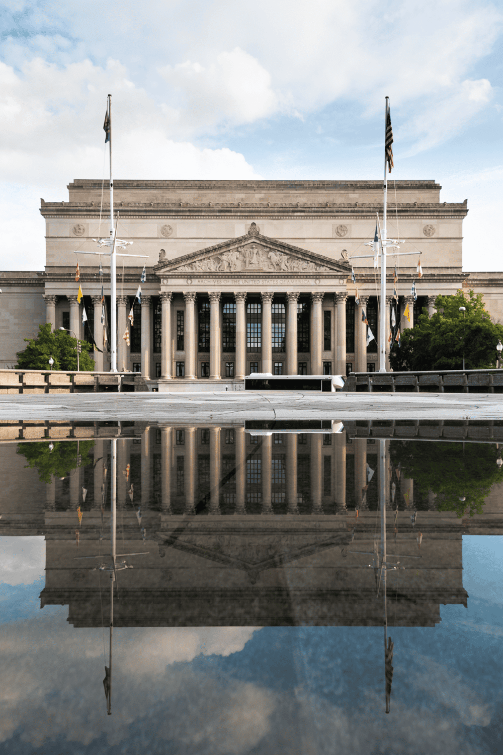 Historic government building with Greek columns, U.S. flags, and reflection on the ground.