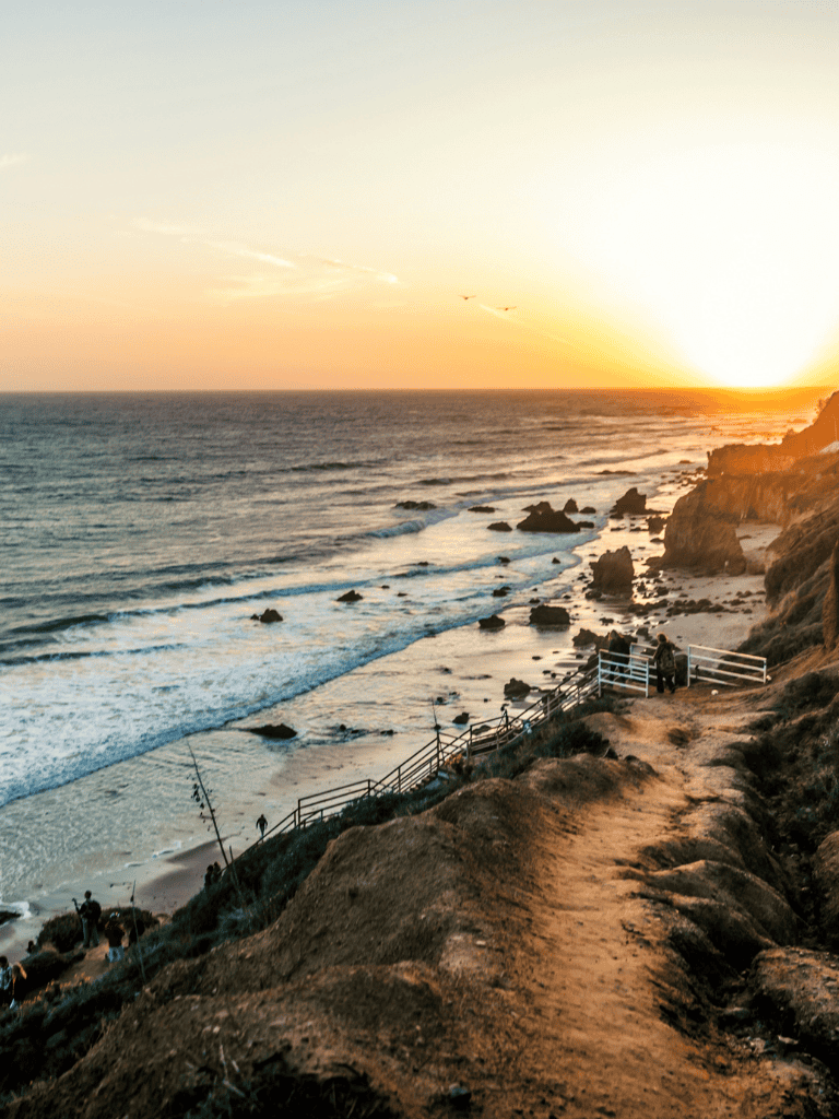 Scenic coastal sunset view with walking trail, rocky shoreline, and ocean waves.