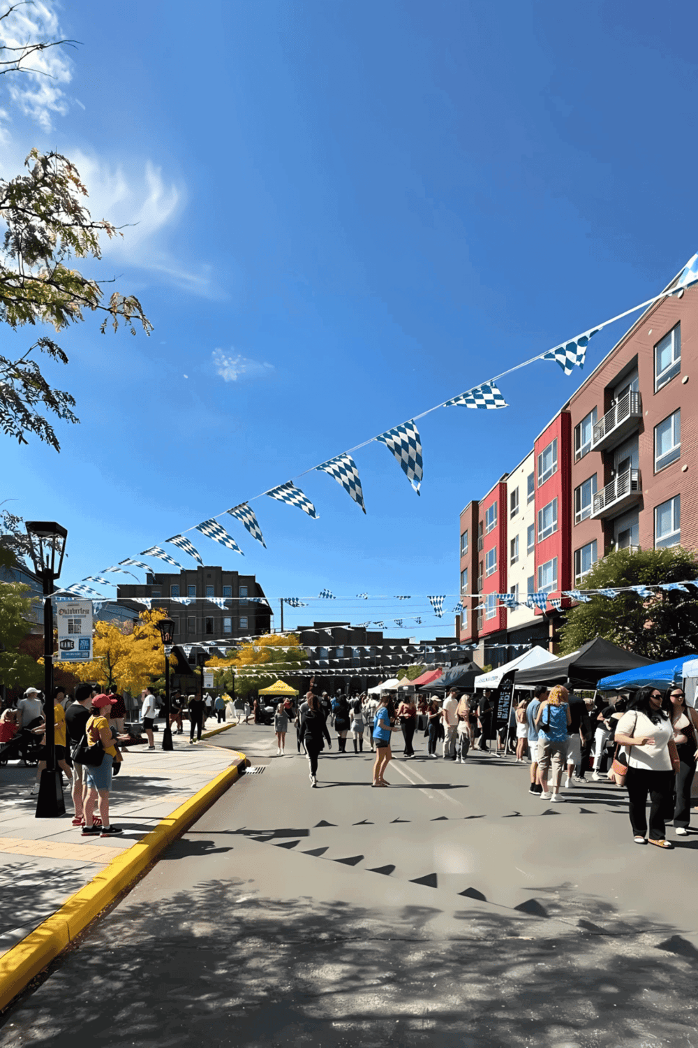 Flags hang above a lively street fair scene with crowds, tents, and colorful buildings under a bright blue sky.