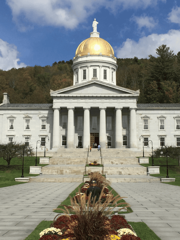 White historic capitol building with gold dome and steps, surrounded by fall foliage.