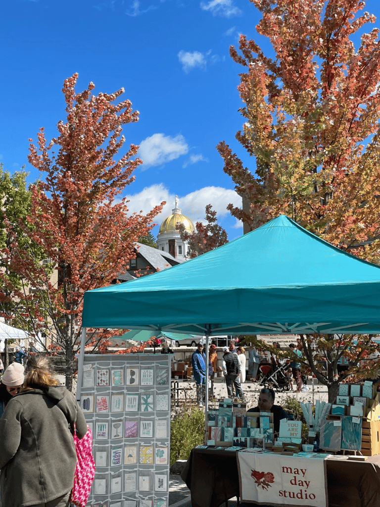 Vibrant outdoor market under fall trees with vendor stalls, shoppers, and a historic dome building in the background.