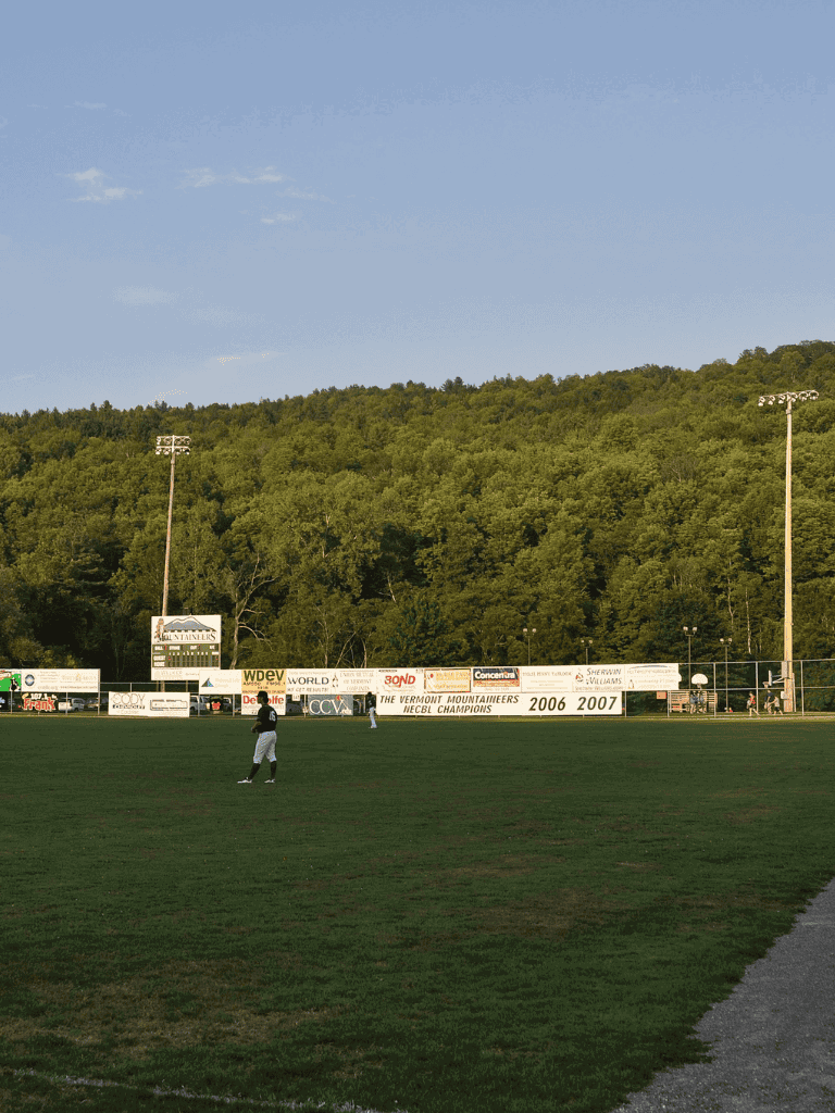Open baseball field with players, lush green hills in the background, and advertisements on the fence.