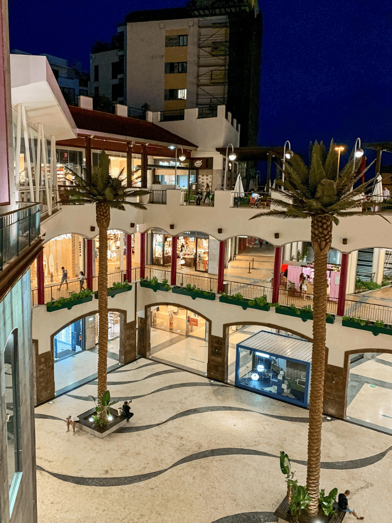 Vibrant downtown shopping mall with outdoor seating, palm trees, and illuminated storefronts at night.