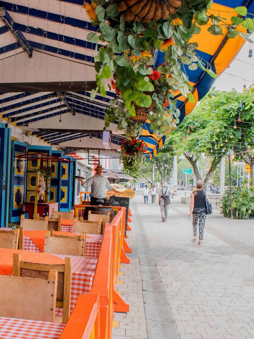 Colorful outdoor restaurant terrace with hanging flower baskets, vibrant umbrellas, and city street scene with pedestrians.