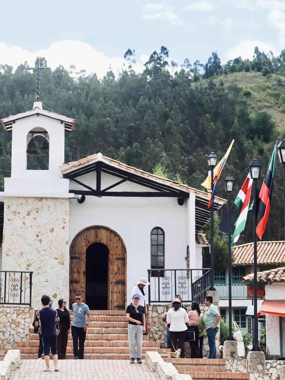 Quaint mountain village church with visitors, colorful flags, and scenic green hills in the background.