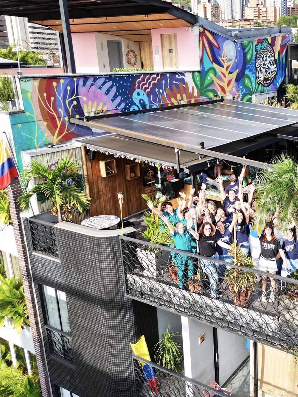 Colorful rooftop bar with mural art, potted plants, and a group of smiling people celebrating.