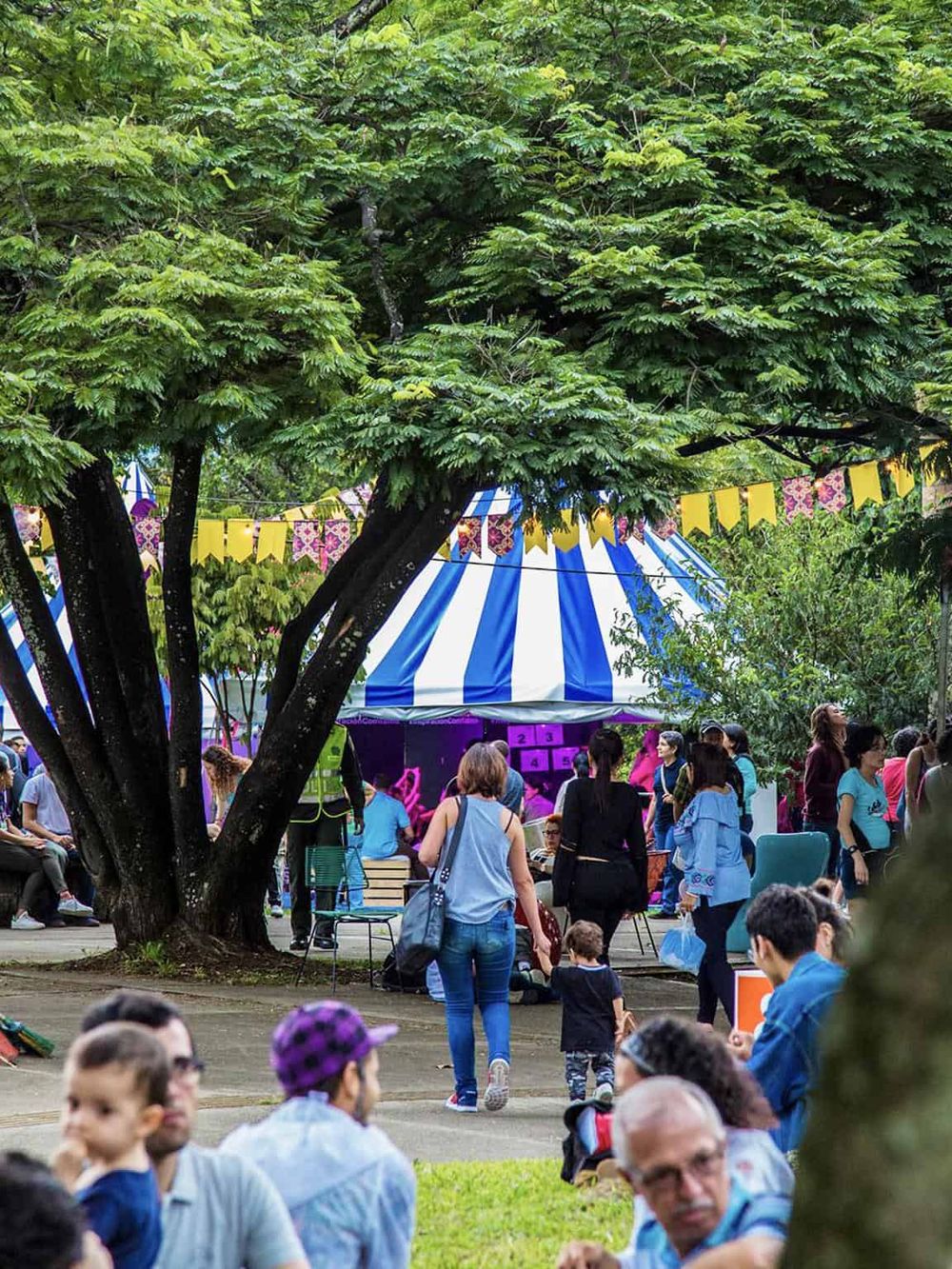 Colorful outdoor event with a striped tent, trees, and attendees enjoying a festival or fair atmosphere.