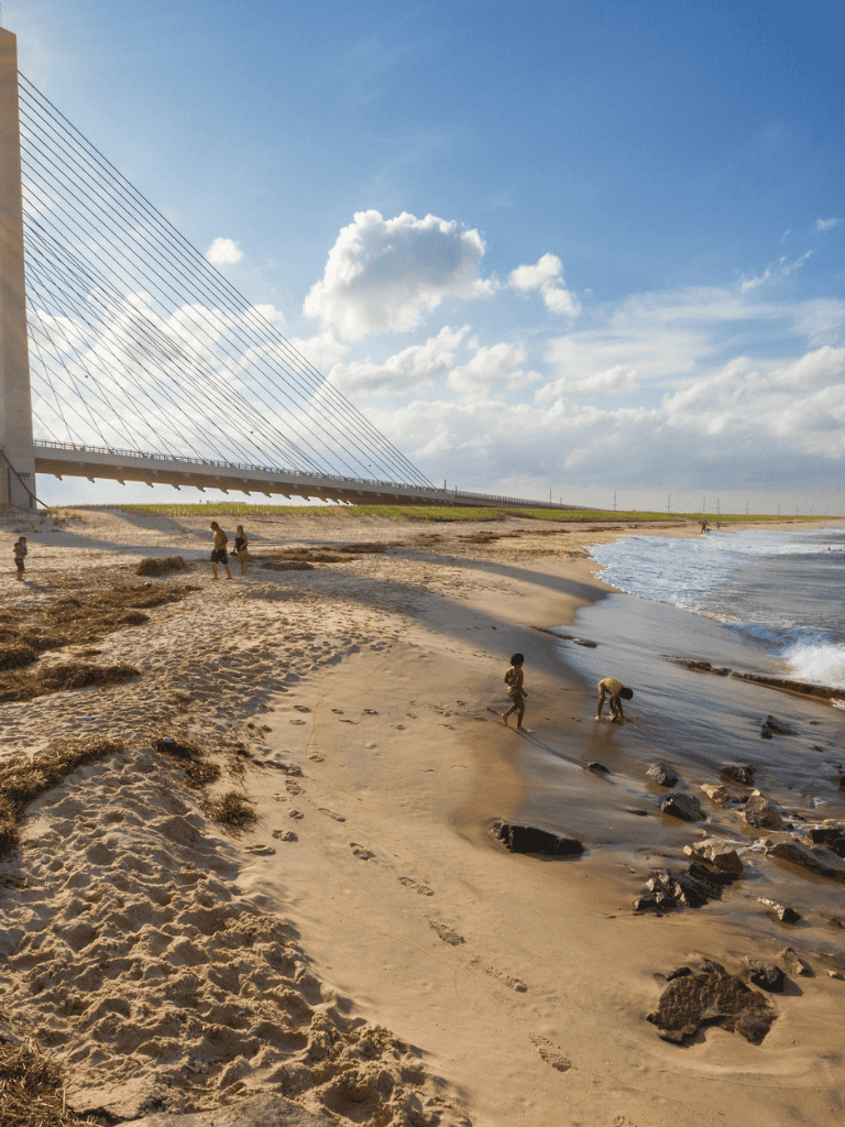 Bright beach scene with children playing, under a clear sky by a modern bridge structure, ideal for travel and outdoor activities.
