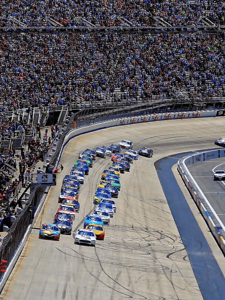 Race cars on a packed stadium track during a NASCAR race, with a large crowd of spectators in the stands.