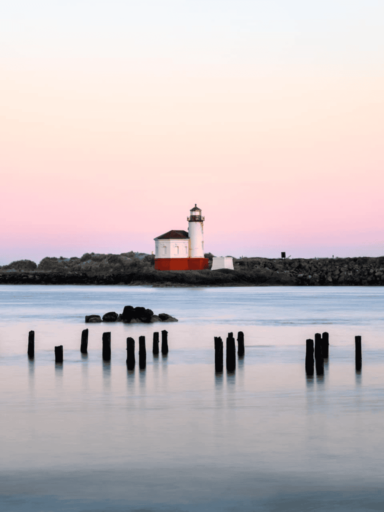 Lighthouse by the water at sunset with calm waves and wooden posts in the foreground.