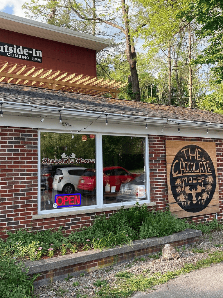 Cozy chocolate shop exterior with "The Chocolate Moose" sign and "Open" neon sign, surrounded by greenery and parking lot.