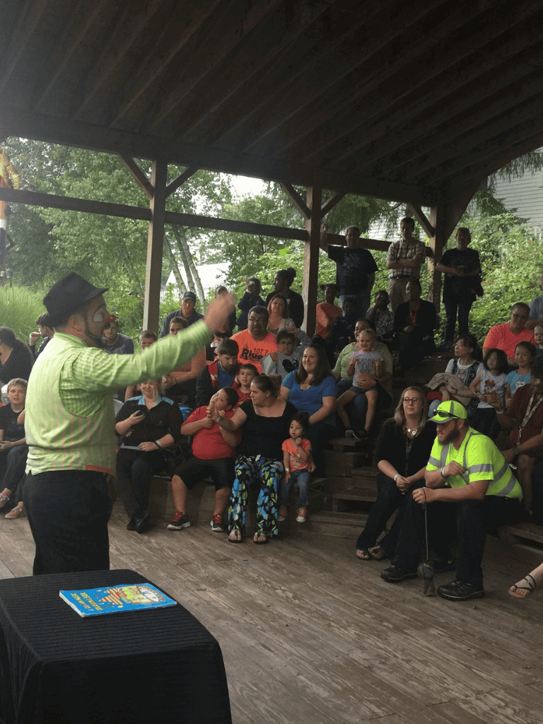 Clown entertaining outdoor audience at community event, kids and adults enjoying live performance on rustic stage.