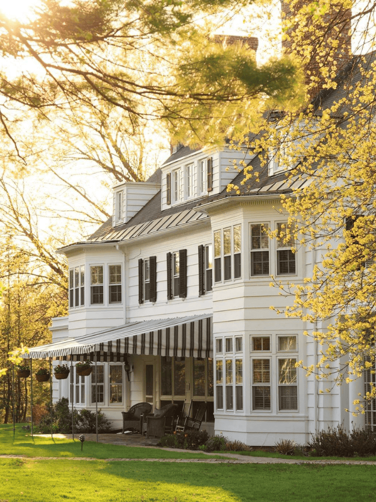 Charming white Victorian house with a striped awning and lush green lawn, surrounded by trees during golden hour.