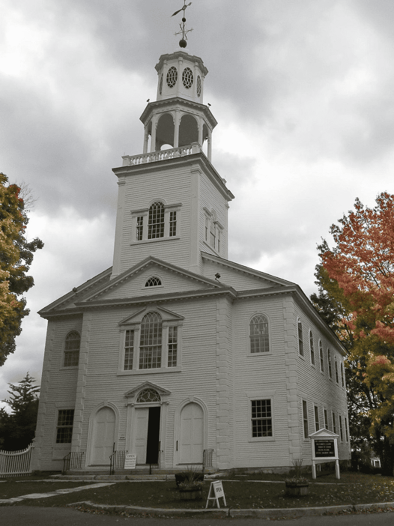 White historic church with tall steeple, fall trees, and cloudy sky.