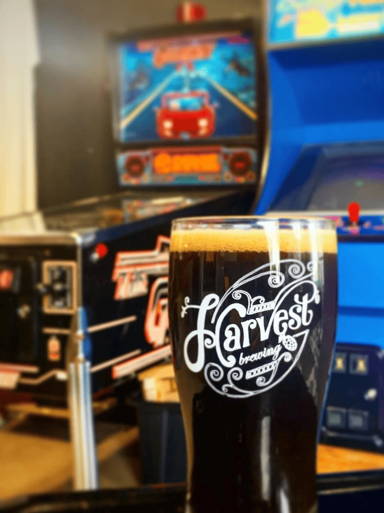 Beer glass with "Harvest Brewing" logo in front of arcade machines and pinball game.