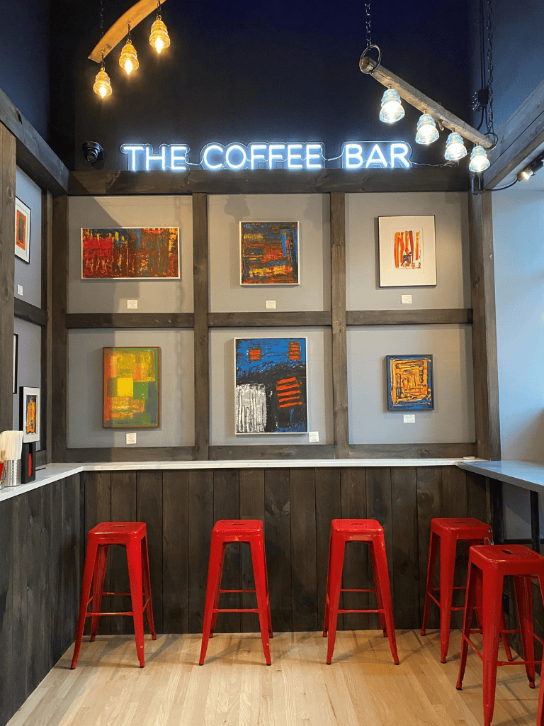 Art gallery coffee bar, vibrant artwork, cozy coffee shop ambiance, modern decor, colorful paintings, seating area, casual cafe atmosphere.