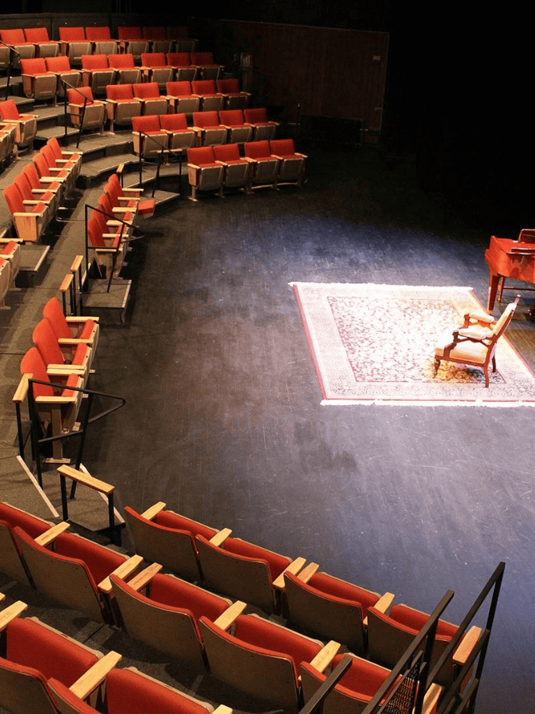 Intimate theater or lecture hall with red cushioned chairs and a stage featuring a reading or speech area.