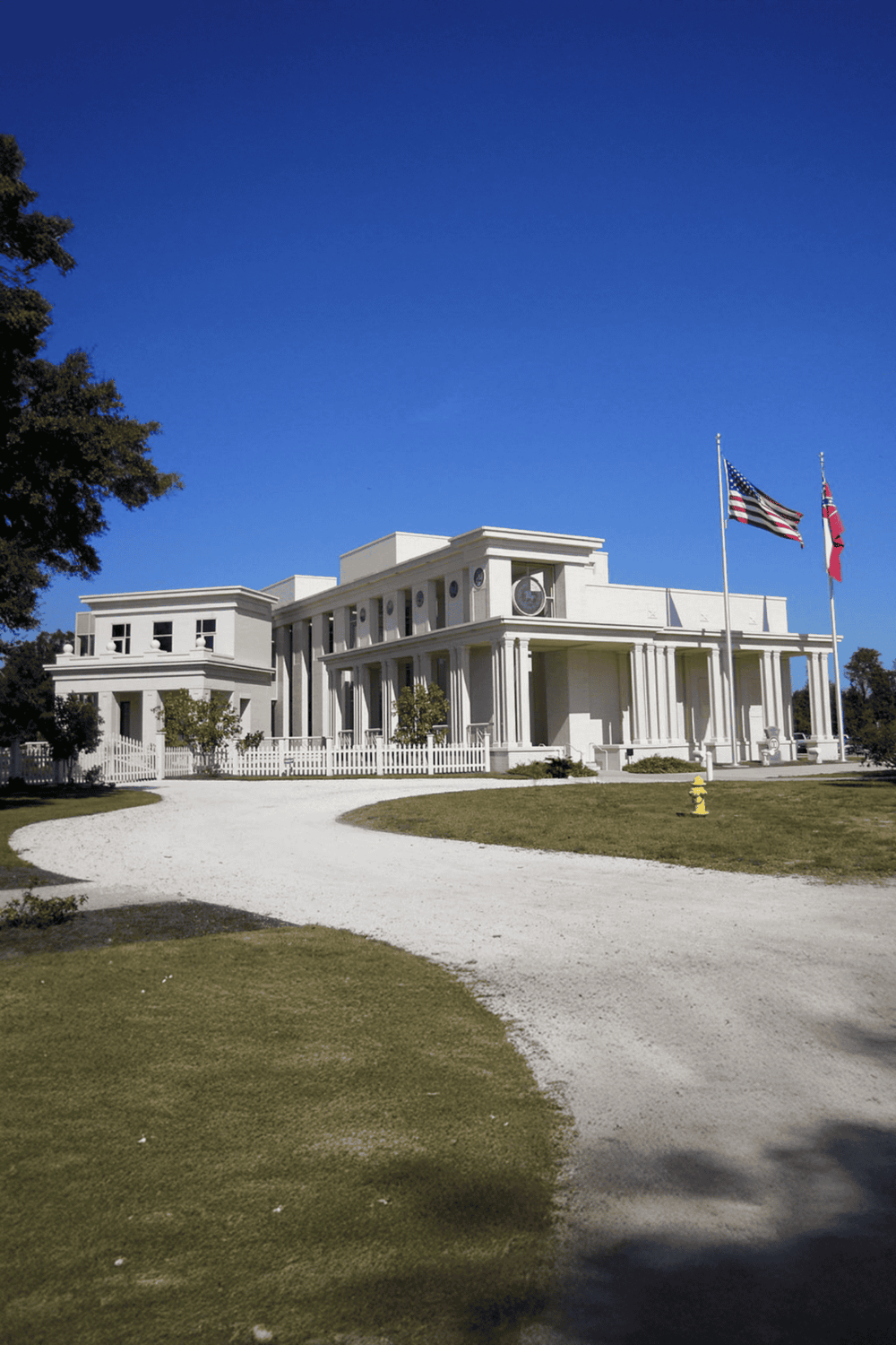 Grand white historic building with American flags, blue sky, and well-maintained lawn.