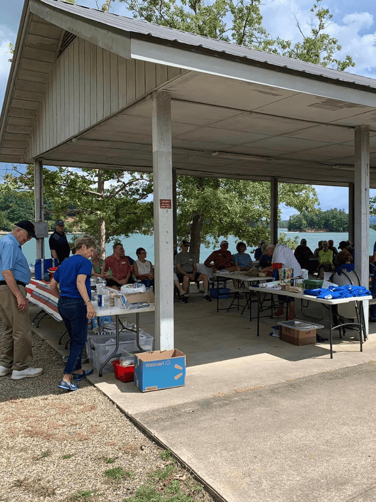 Community outdoor gathering under pavilion by water with food and supplies, people enjoying a sunny day.