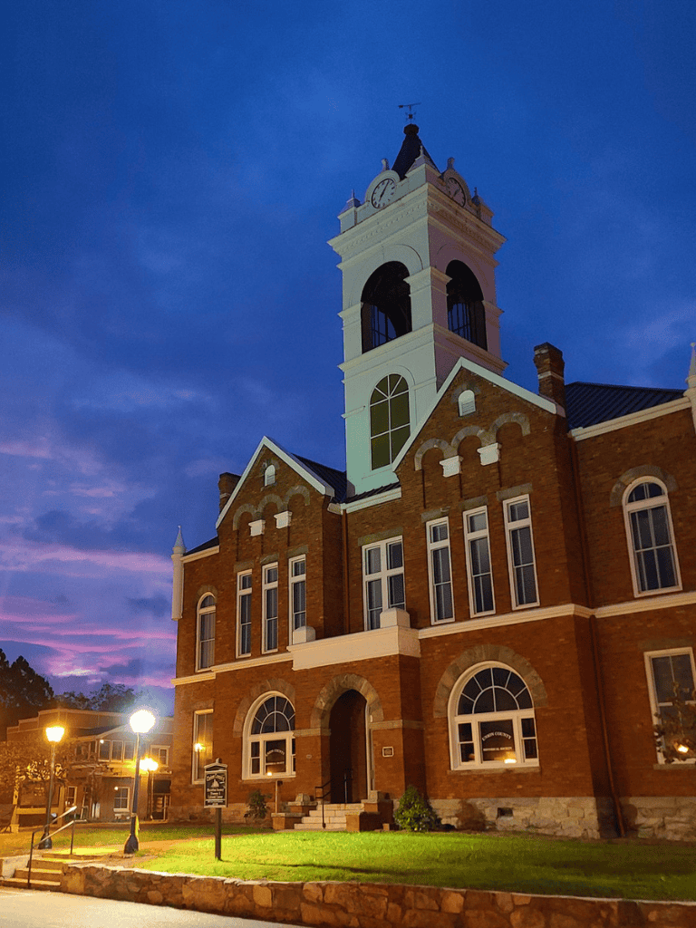 Clock tower on historic brick courthouse at dusk, illuminated by streetlights.