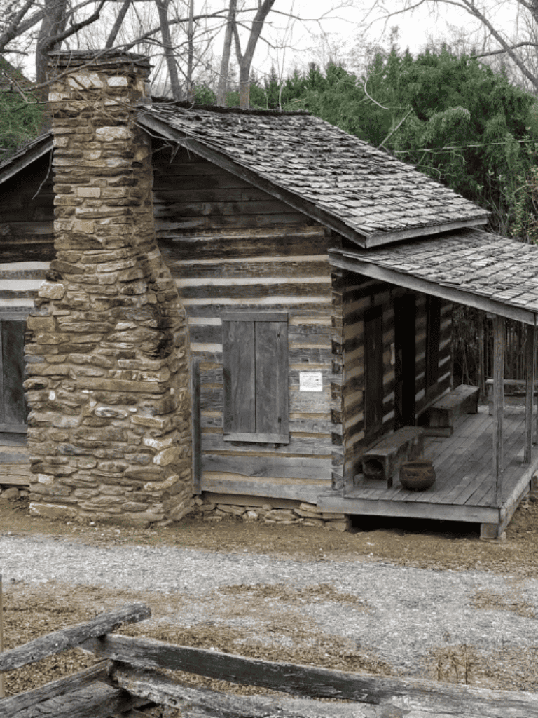 Rustic wooden cabin with stone chimney and porch in historical outdoor setting.