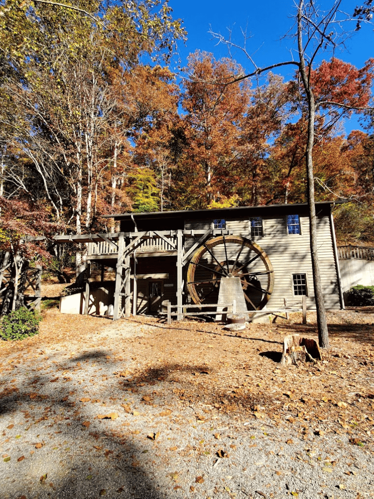 Rustic watermill in a scenic autumn forest setting with colorful trees and clear blue sky.