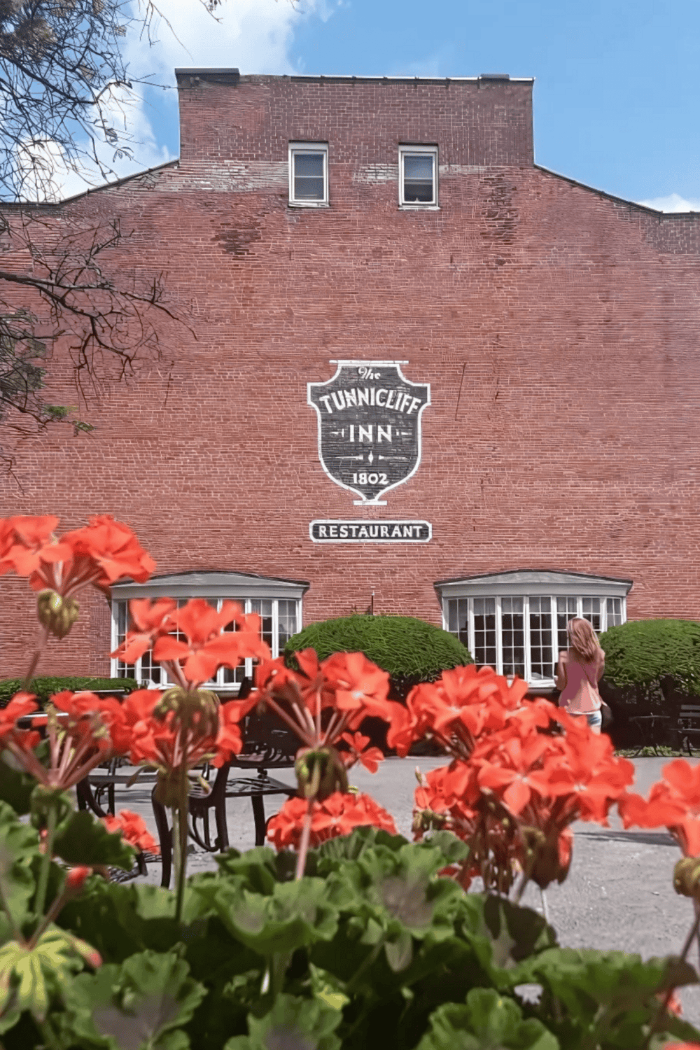 Old brick hotel exterior with "Tunnicliffe Inn" sign, lush greenery, and a woman on a sunny day.