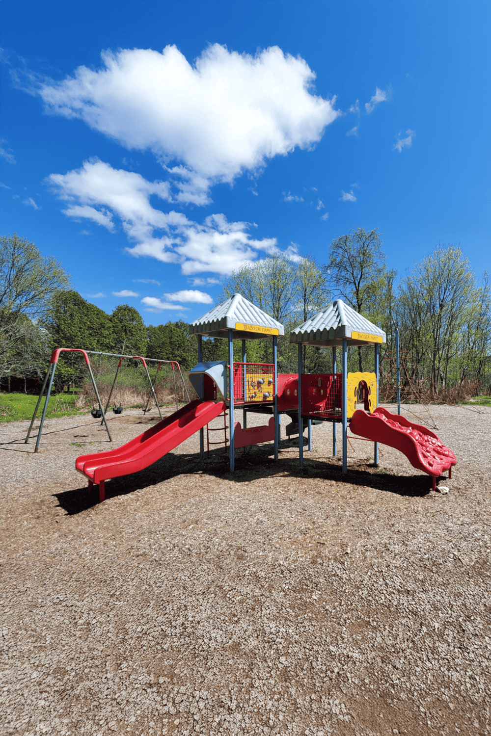 Bright red playground slides and swings at Badger Park under clear blue sky with fluffy clouds, ideal for family fun and outdoor activities.