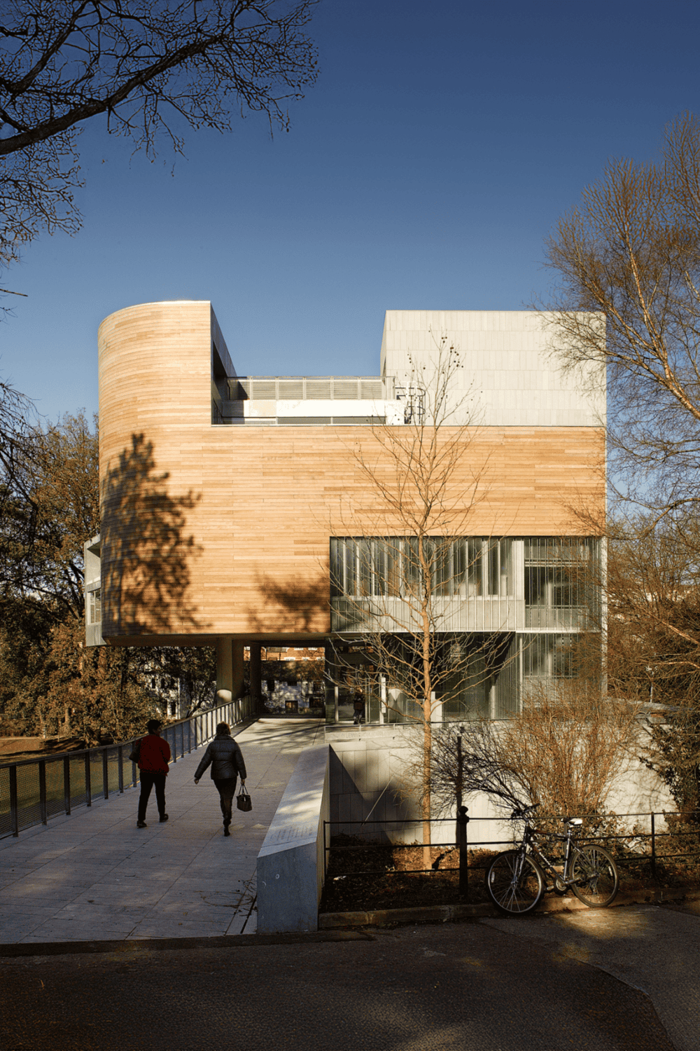 Contemporary modern architecture building with wooden and glass elements, park surroundings, and pedestrians.