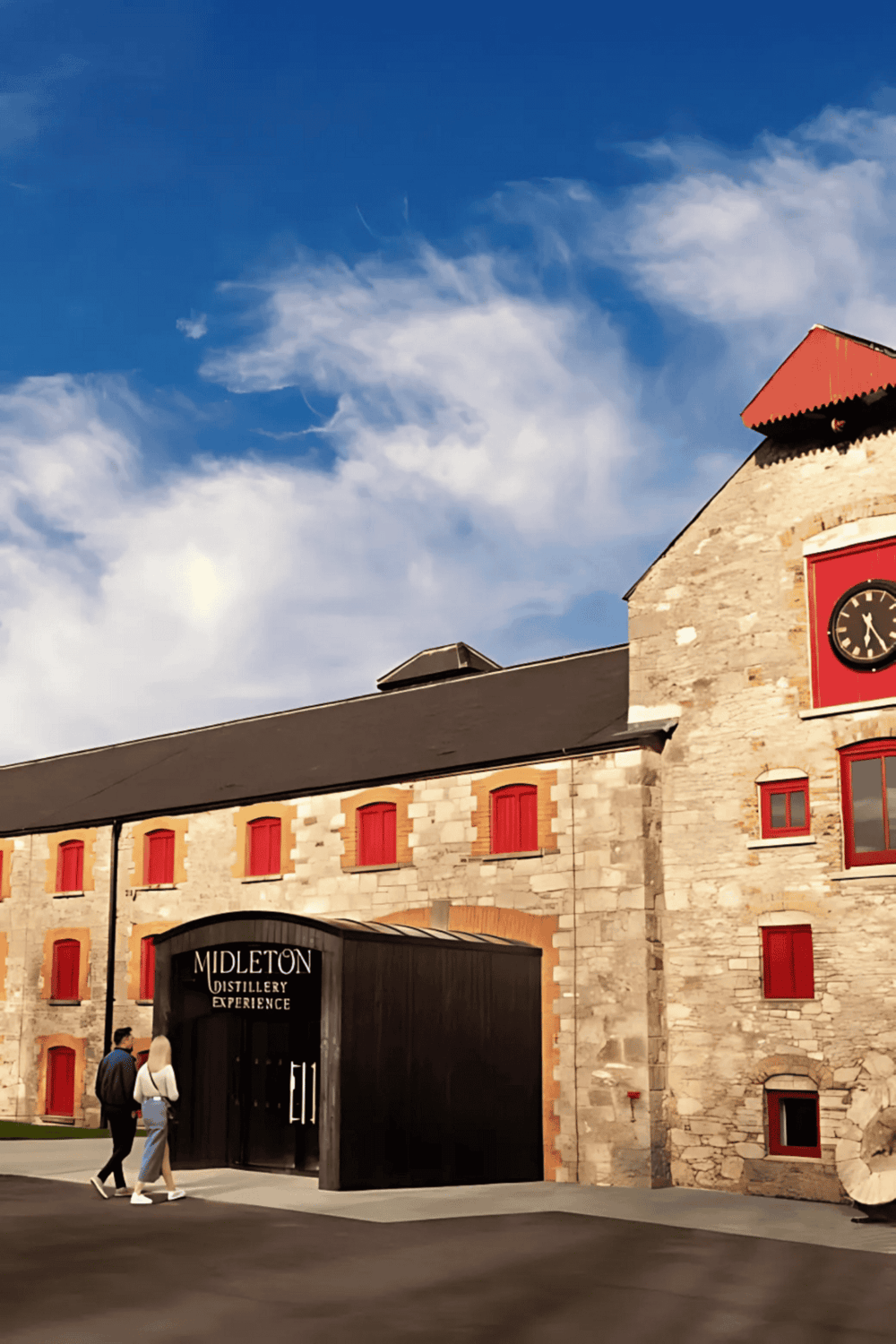 Historic distillery building with red window shutters and a modern entrance, under a bright blue sky.