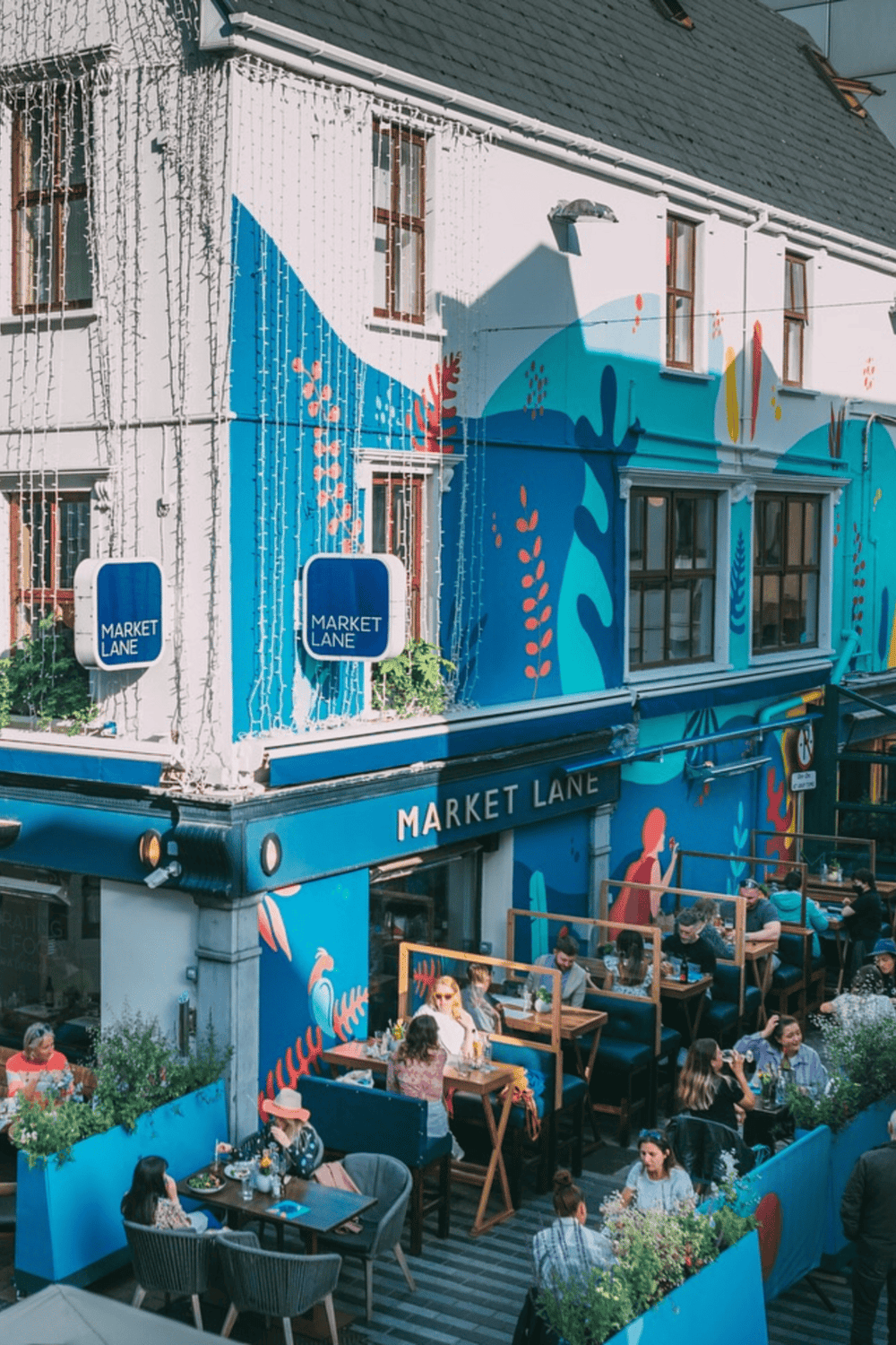 Colorful mural on building exterior with outdoor dining area in an urban setting, promoting travel and local cuisine.