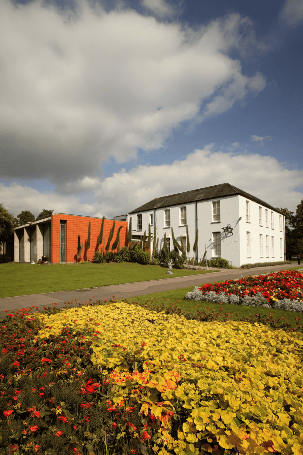 Colorful garden with historic white building and modern red structure under blue sky, showcasing architectural and landscaping design.