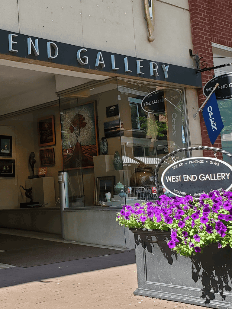 Colorful art gallery storefront with flower planters and "Open" sign on busy street.