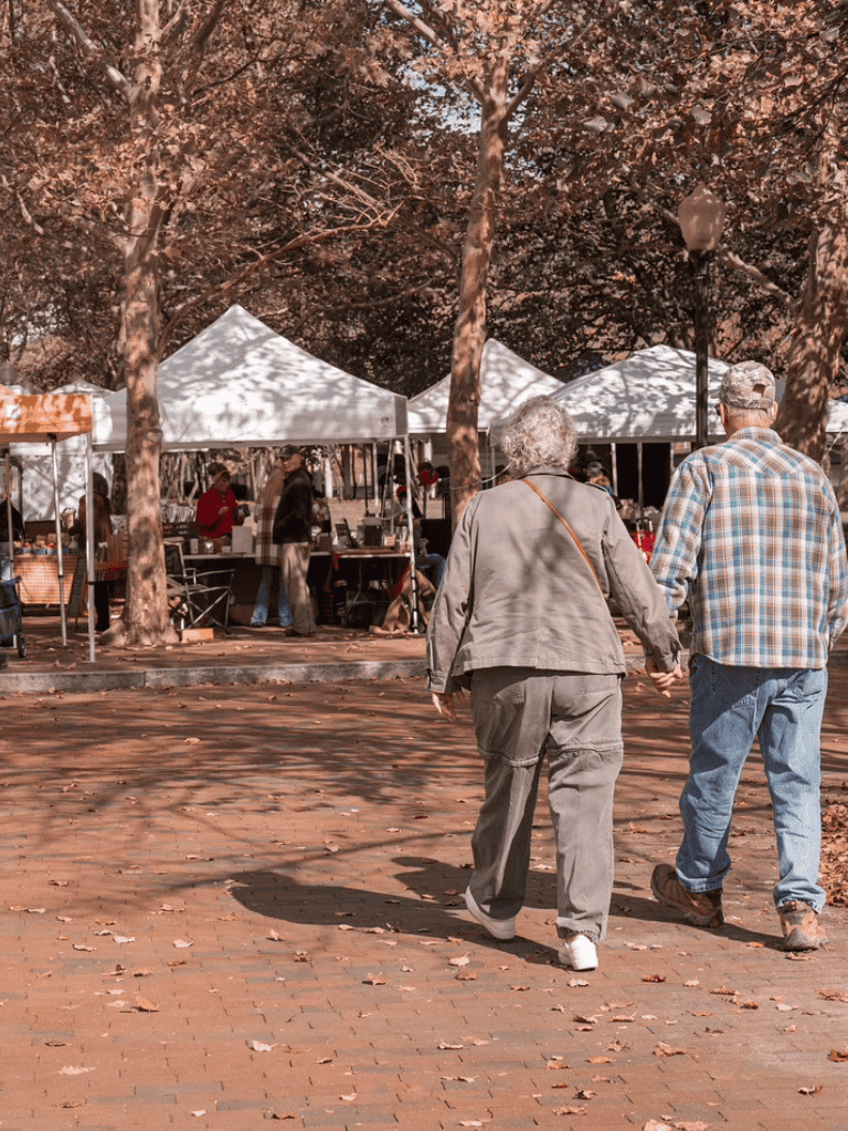 Colorful outdoor market with tents and shoppers, autumn trees, senior couple walking hand-in-hand.