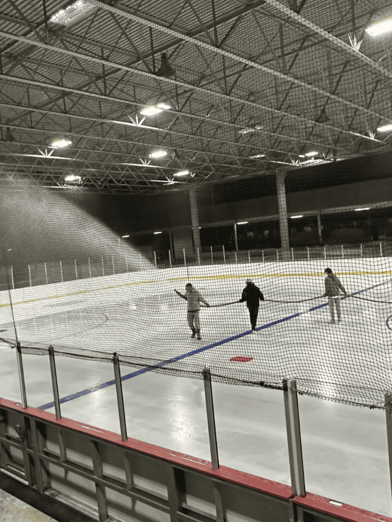Ice skating rink with three people holding hands, under a large industrial ceiling.