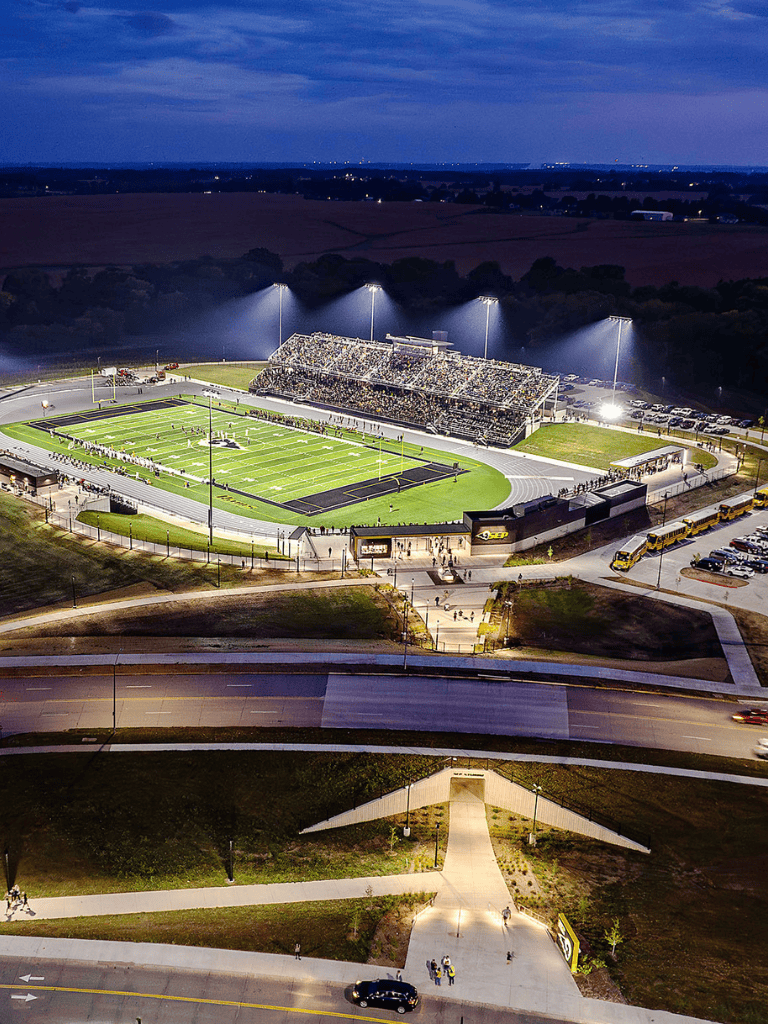 Brightly lit football stadium at dusk with seating, field, and surrounding roads.