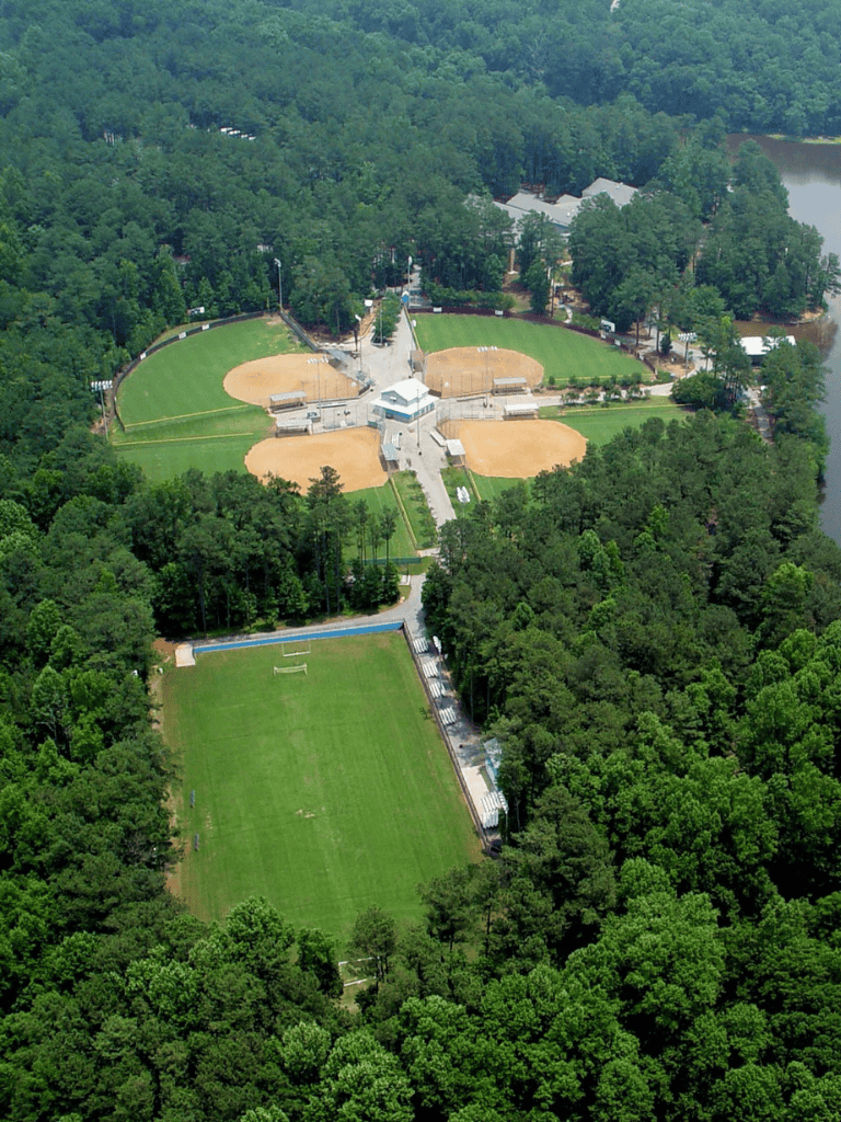 Aerial view of Quest for Directions sports complex surrounded by lush green trees and a lake in the distance.