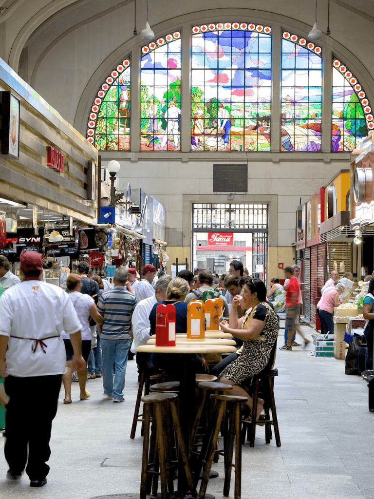 Colorful stained glass window at indoor market with people dining and shopping, vibrant atmosphere and diverse cuisine.