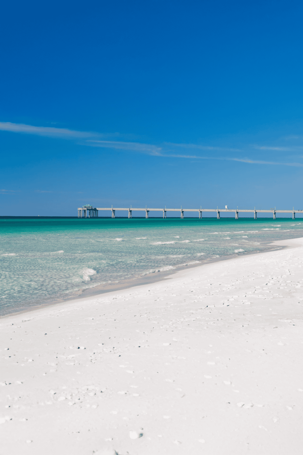 Seaside pier extending into clear turquoise ocean with white sandy beach and bright blue sky.