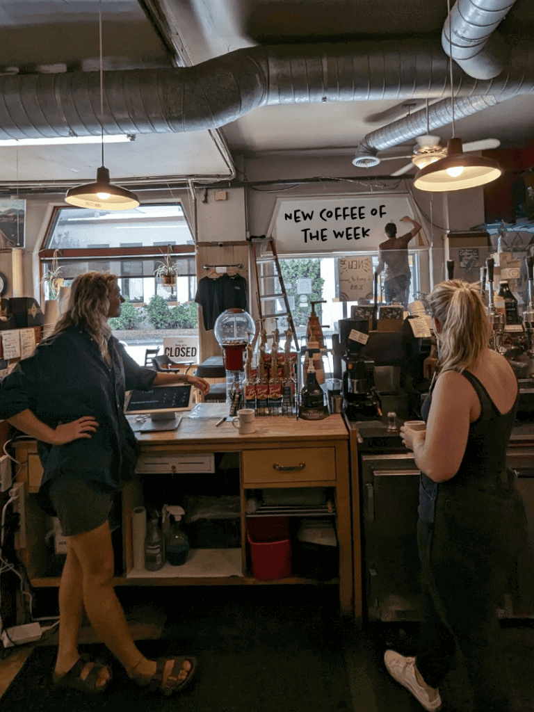 Cozy coffee shop interior with baristas, modern industrial decor, and a sign reading "New Coffee of the Week" for a welcoming cafe experience.