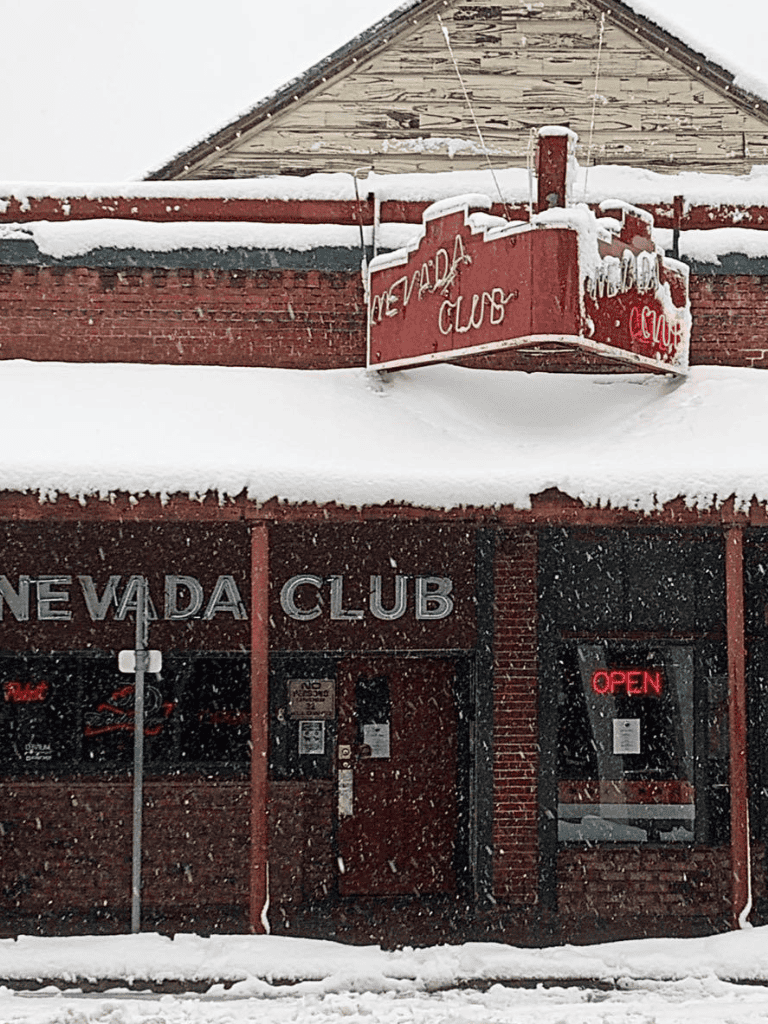 Snow-covered Nevada Club building in winter, with neon "Open" sign visible.