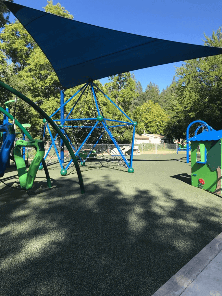 Colorful outdoor playground equipment at a park with shade sail and green trees.