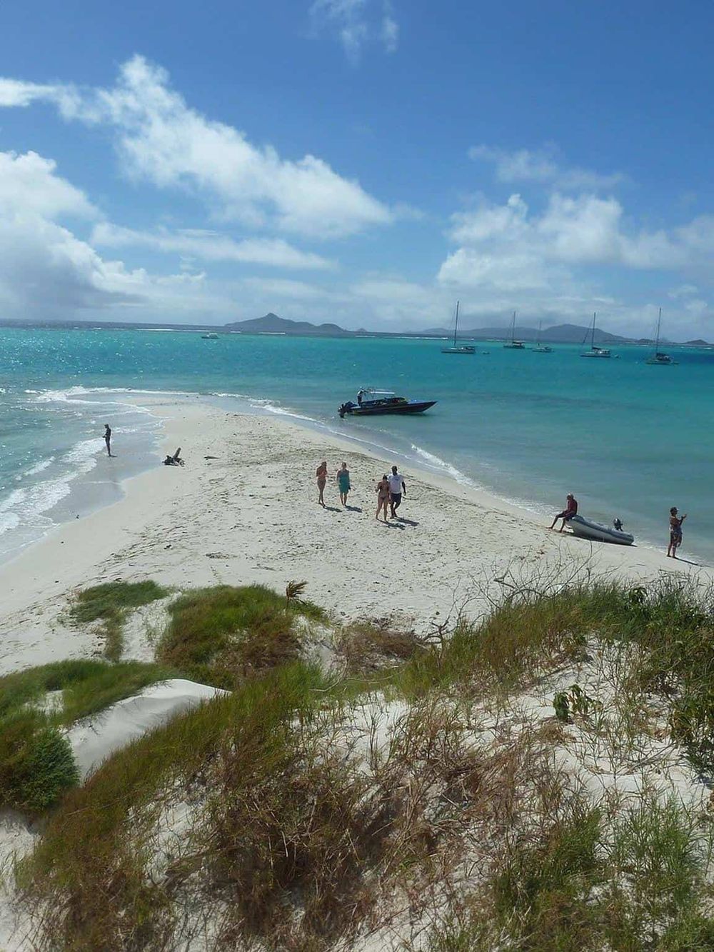 Secluded tropical beach with white sand and turquoise waters, boats anchored offshore, and people enjoying the ocean scenery.