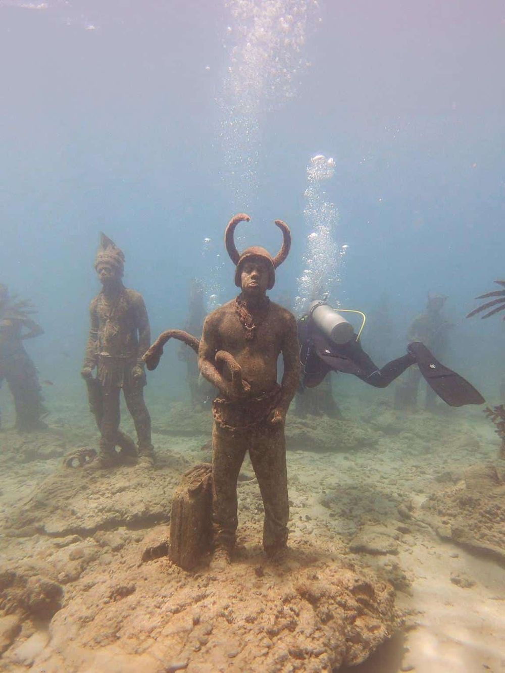 Underwater statues depicting tribal figures in a boat in a clear ocean.