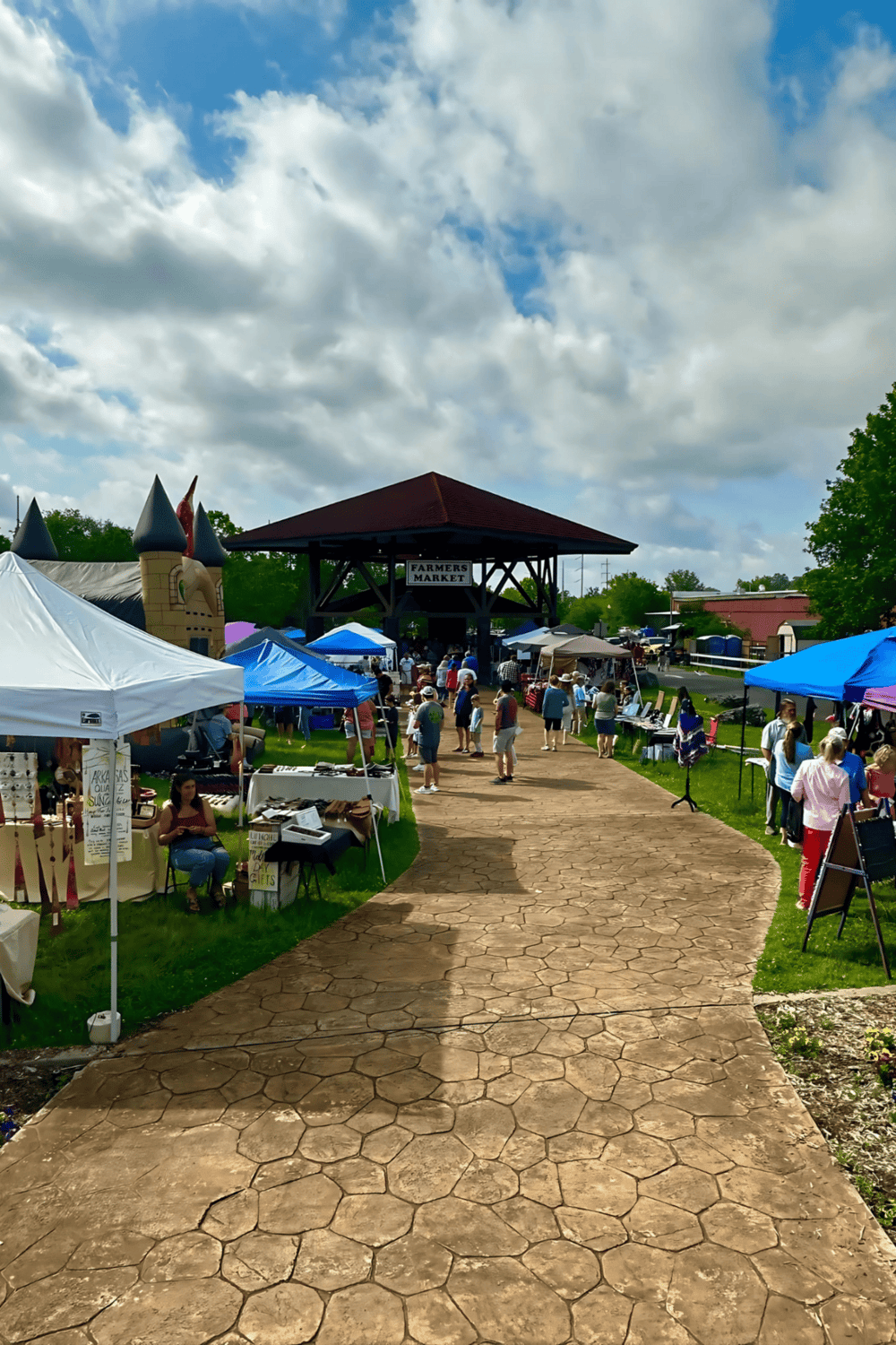 Colorful farmers market with vendor tents and shoppers under partly cloudy sky.