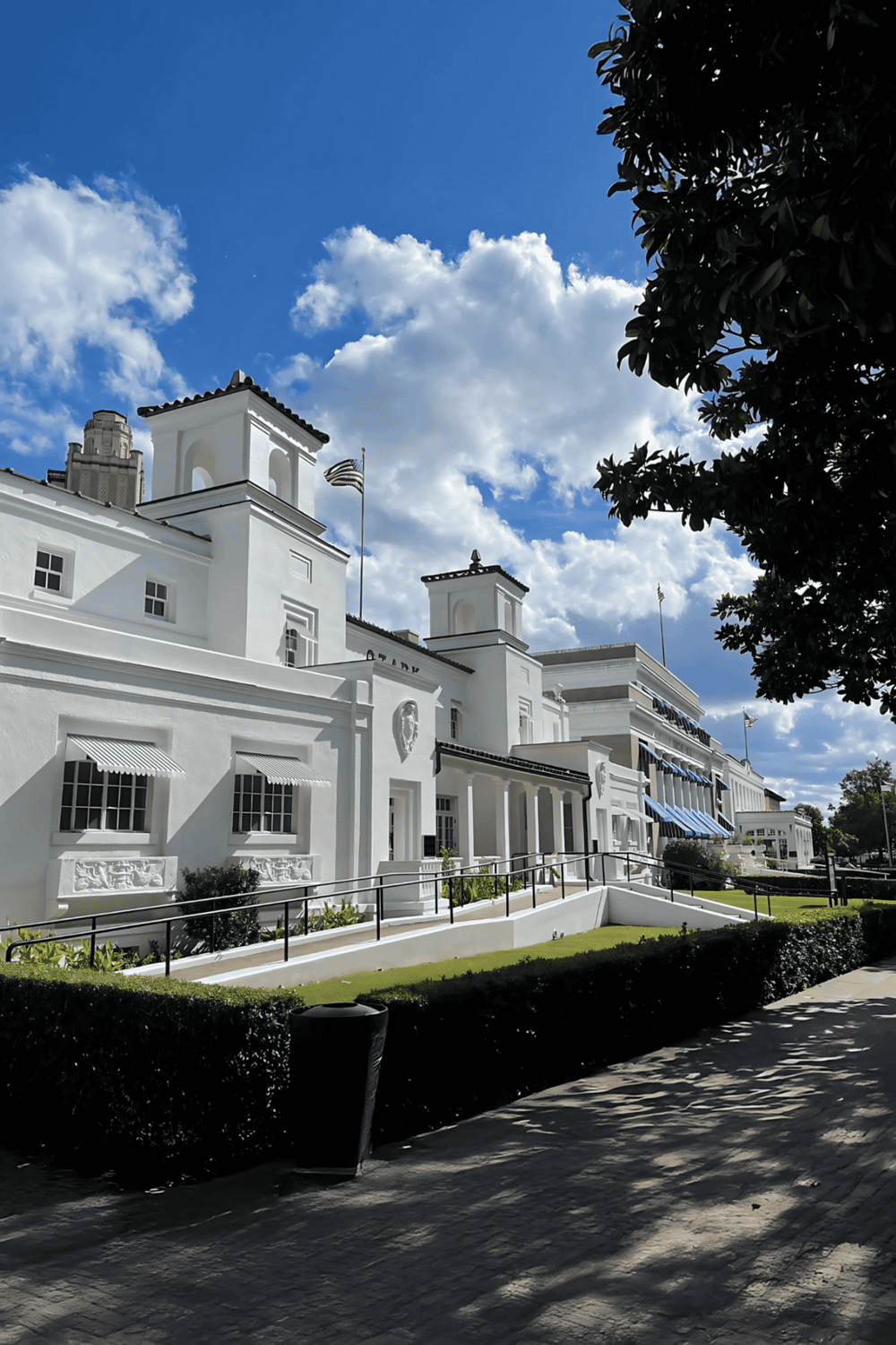 White historic building with blue sky and clouds, featuring classic architecture and lush greenery.