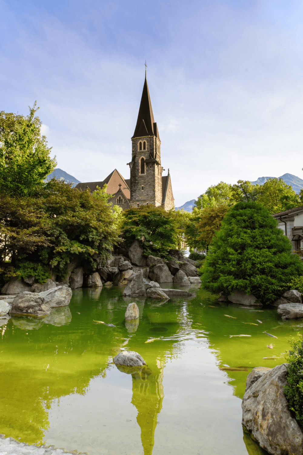 Bright church with tall steeple overlooking a pond with koi fish and lush greenery in a scenic mountain town.